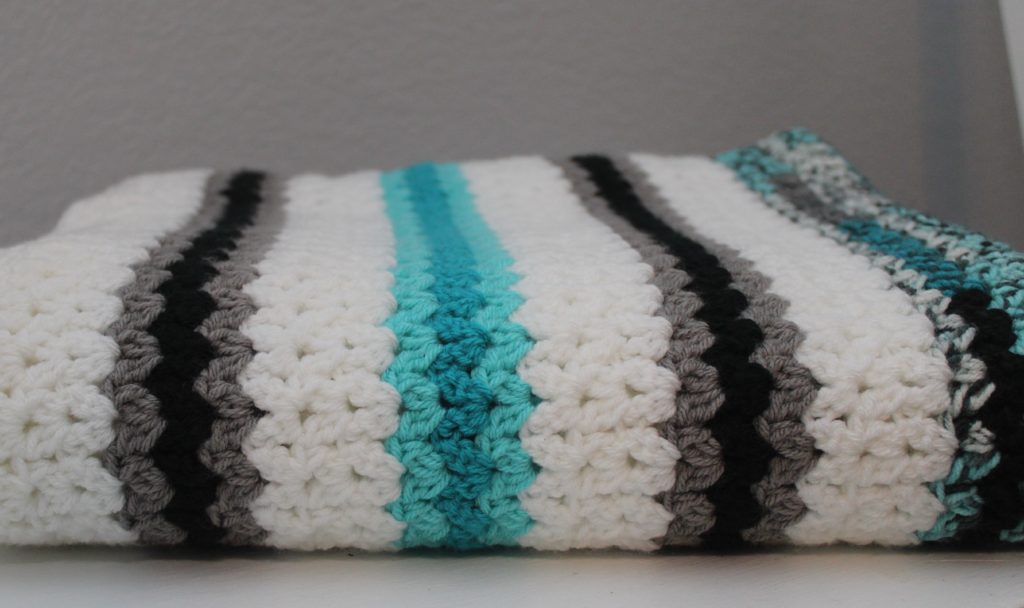 Beautiful and colorful cluster v-stitch throw in teal, grey, white, and black draped over grey chair.