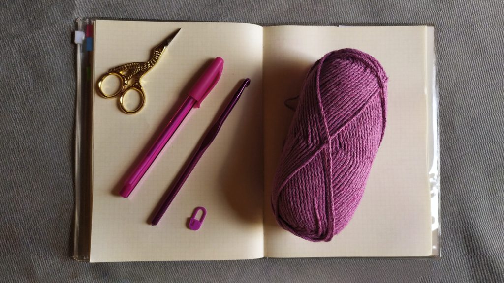 yarn, crochet hook, pen, and scissors laying on an open journal
