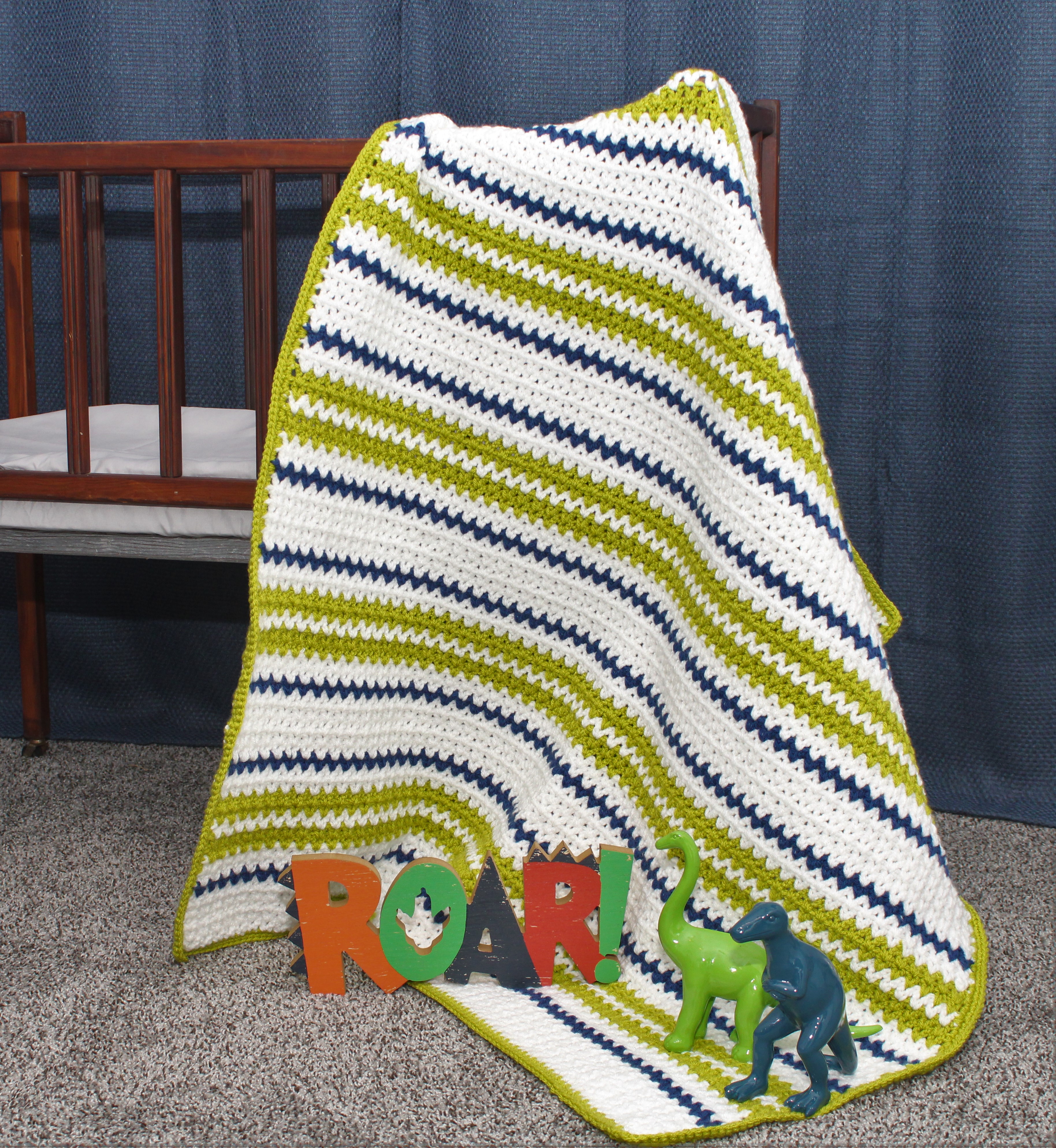 white gren and blue baby afghan draped over a crib with blue and green dinosaurs on the edge with the word roar!