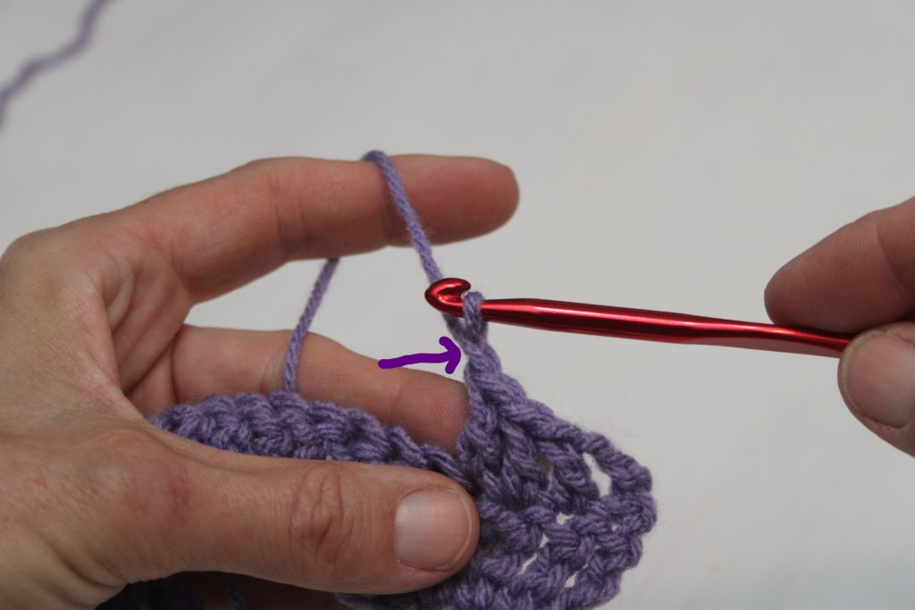 hands hold purple yarn and a red crochet hook showing a connecting chain
