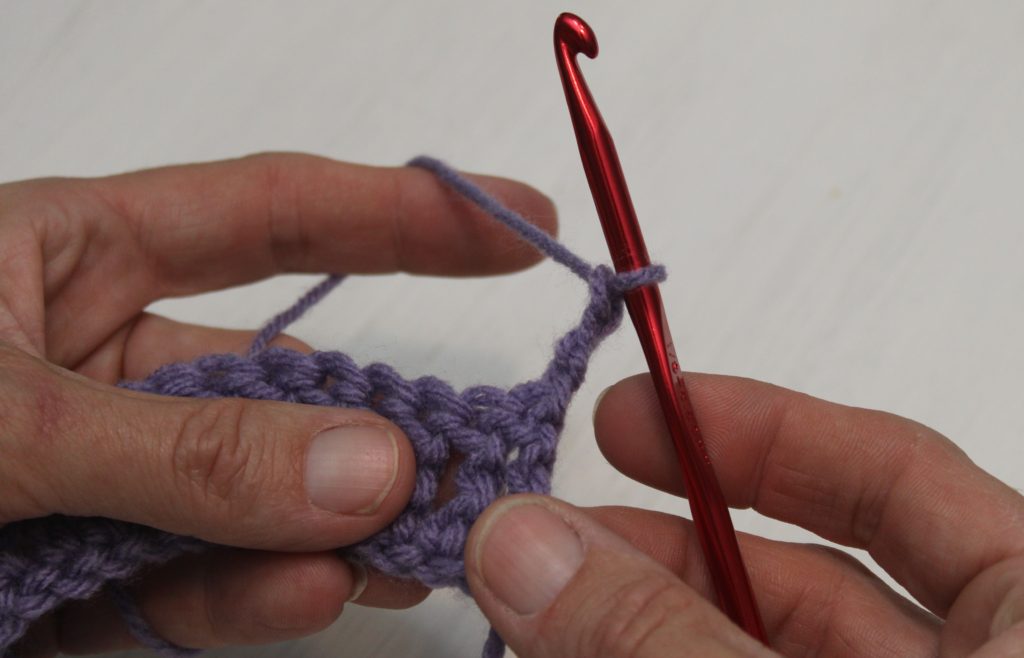 hands holding purple yarn and a red crochet hook, showing a turning chain