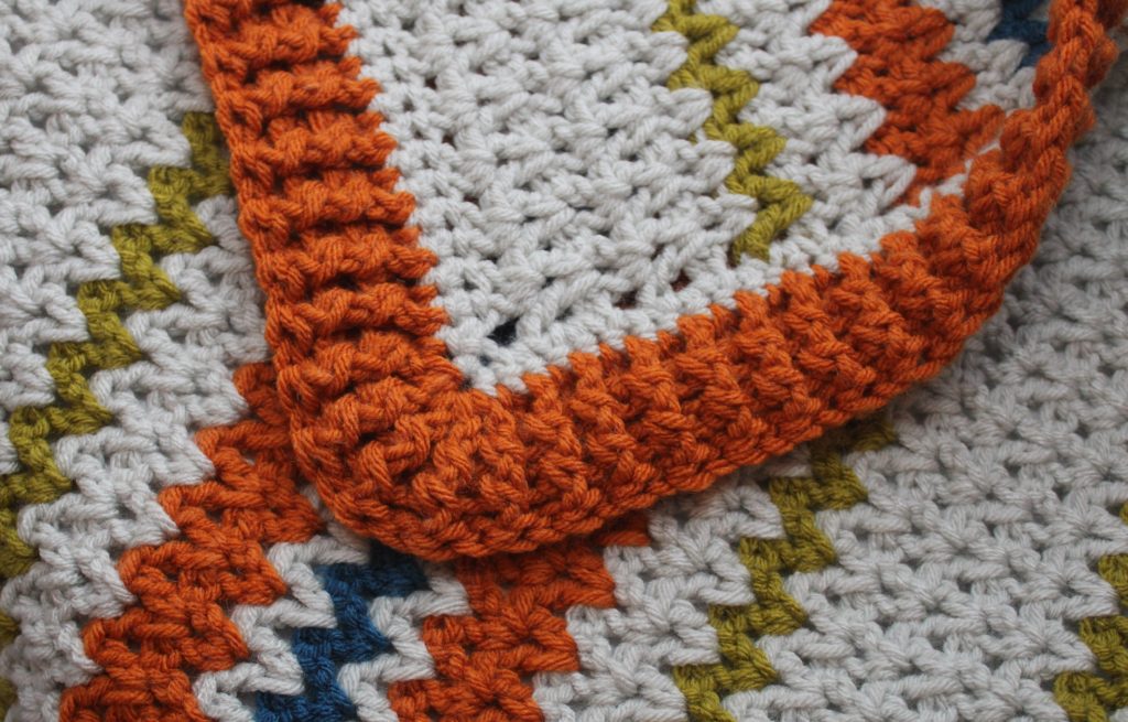 Crocheted blanket in orange, grey, blue and green showing the border.