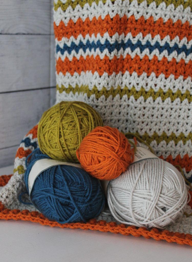 work zone baby blanket in orange, blue, green and grey balls of yarn sitting on a blanket draped over a basket.
