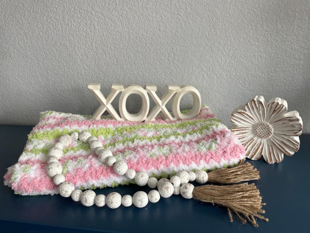 folded  cozy and warm pipsqueak baby afghan in pink, green, and white baby afghan folded and laying on dresser with beads, daisy, and xoxoxo