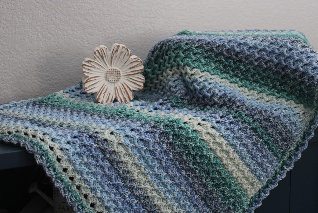 Seahore by Moonlight afghan draped over a table with a daisy, in blues and greens