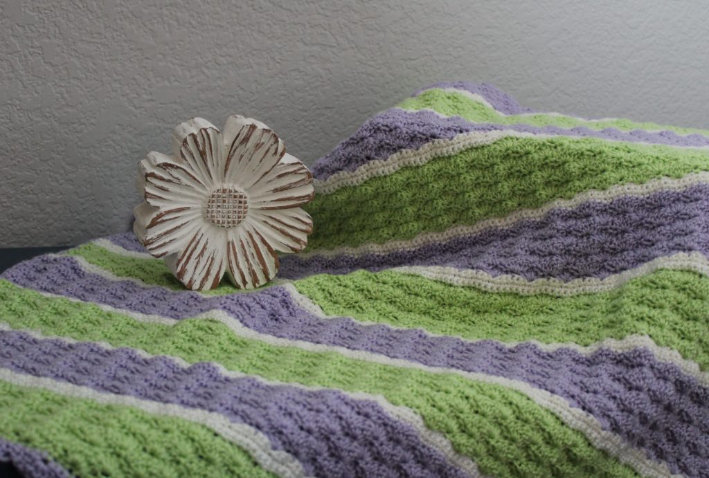 fields of clover  in green, purple, and white shell stitch afghan draped across dresser with a daisy