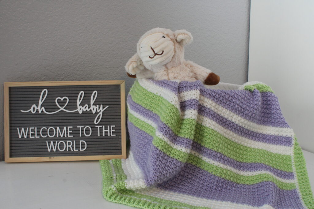 Delightful Springtime Modern Stripe baby afghan in green. white, andpurple draped over a basket with a stuffed lamb