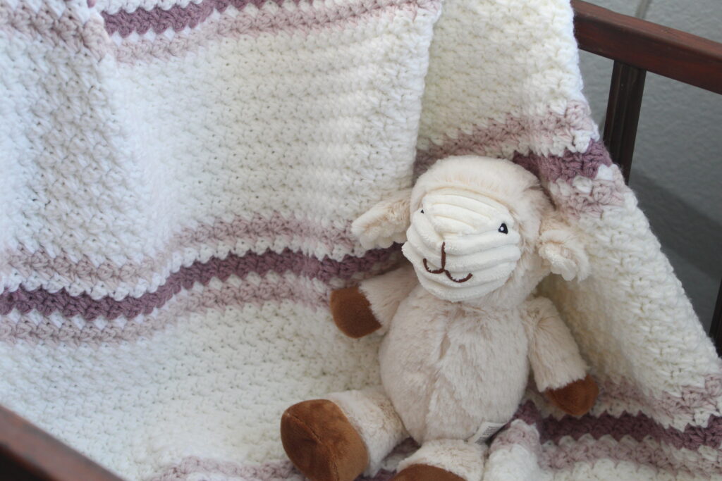 Easy Sugarplum Princess Baby afghan in White crcheted afghan with two tone pink stripes draped over a crib with a stuffed lamb